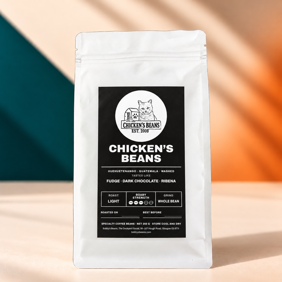 Chicken's Beans coffee with the final black cat label on a white bag, photographed on a beige surface with a teal and orange background.