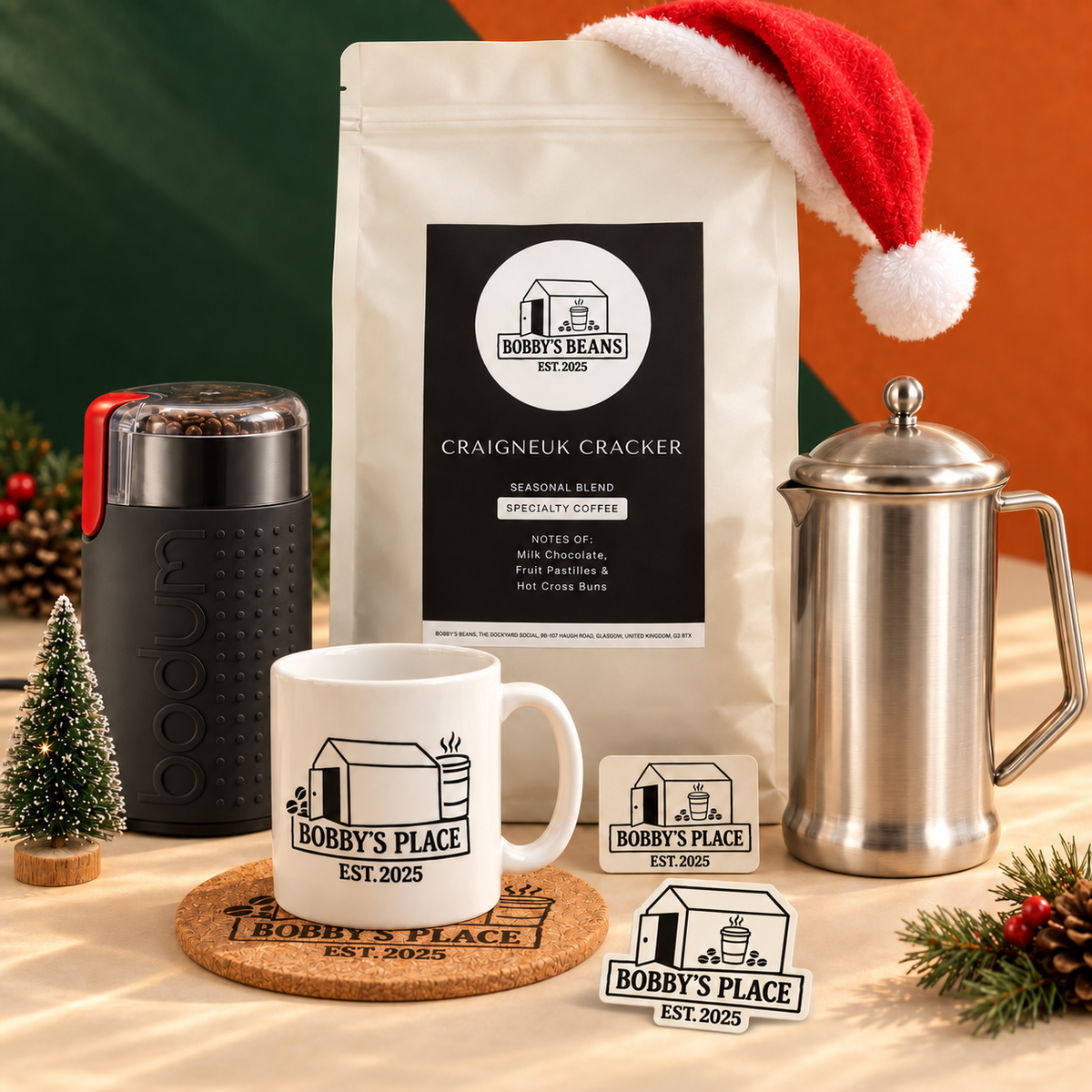 The Christmas Cracker gift set with a 1kg Craigneuk Cracker coffee bag, black Bodum grinder, stainless steel cafetière, Bobby’s Place mug on a large cork coaster and a fridge magnet, styled for Christmas. Includes a Bobby's Place glow in the dark sticker.