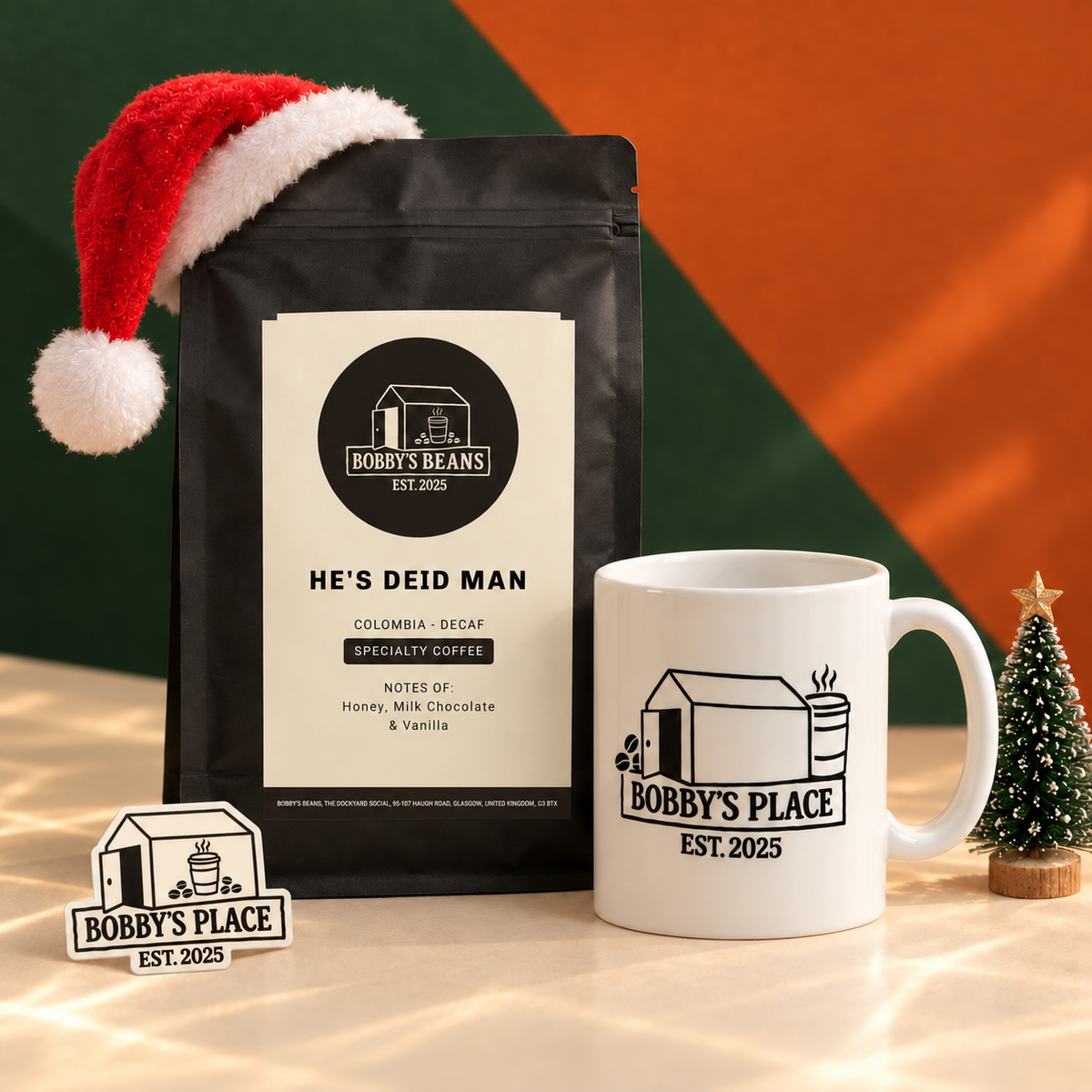 The Nightcap with a black He's Deid Man decaf coffee bag and a white Bobby's Place mug, styled with a Santa hat and miniature Christmas tree. Includes a Bobby's Place glow in the dark sticker.
