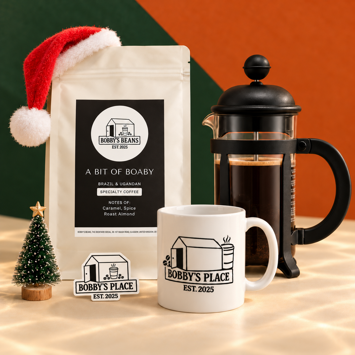 The Starter Kit with a black Bodum Java 3 cup French press, A Bit of Boaby coffee and a Bobby's Place mug, styled with a Santa hat and miniature Christmas tree. Includes a Bobby's Place glow in the dark sticker.